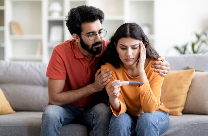 Infertility Problems. Portrait Of Upset Young Indian Couple Looking At Negative Pregnancy Test While Sitting Together On Couch At Home, Caring Eastern Husband Comforting Depressed Wife, Free Space