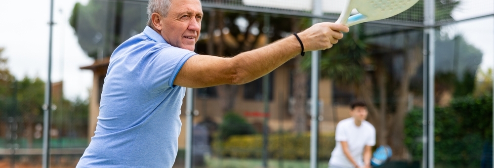 Focused aged man playing friendly paddleball match on outdoor summer court. Senior people sports concept..